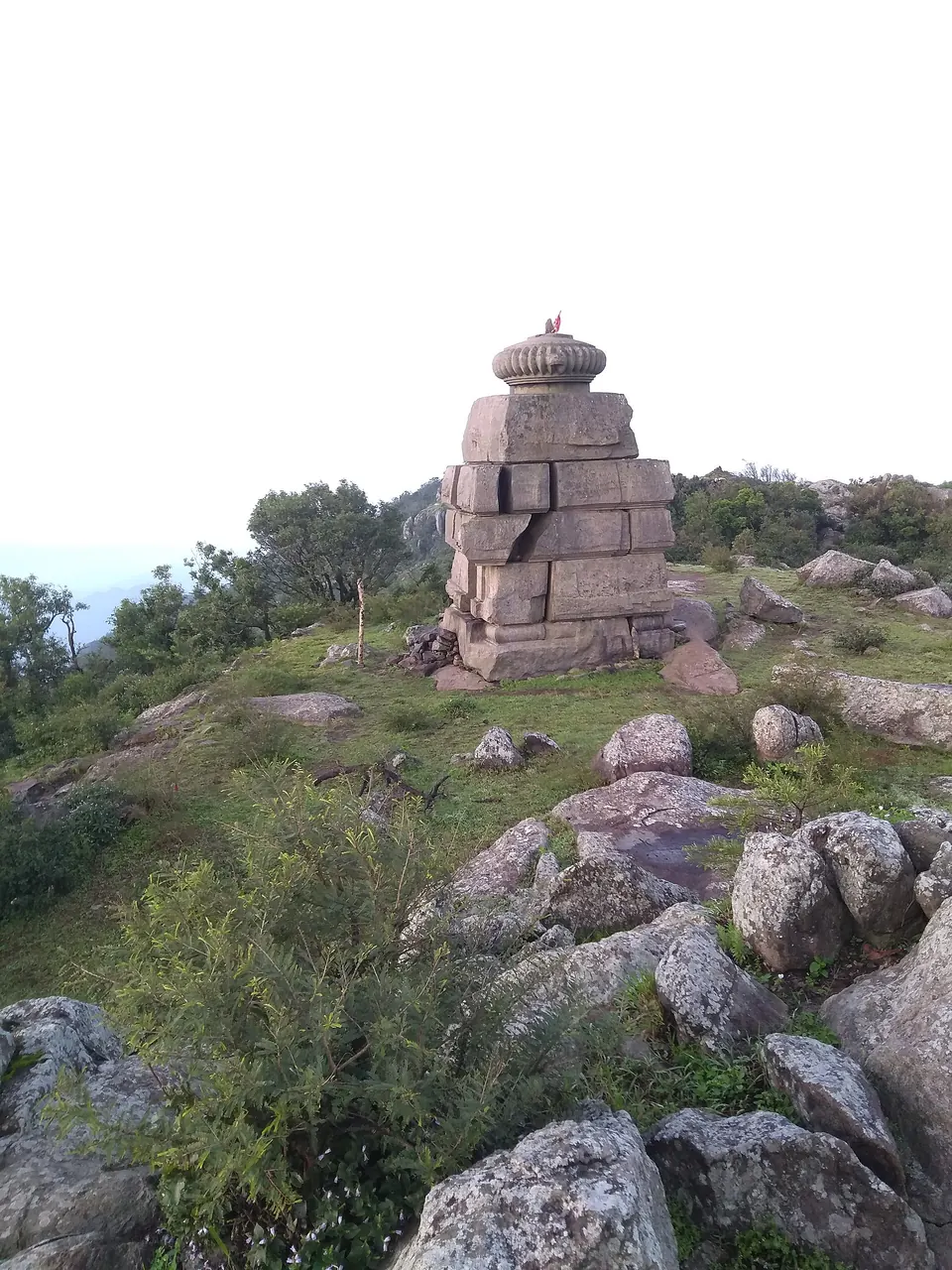 A small square stone shrine built of large grey granite blocks with a round stone top, standing among boulders and bushes on a hilltop.
