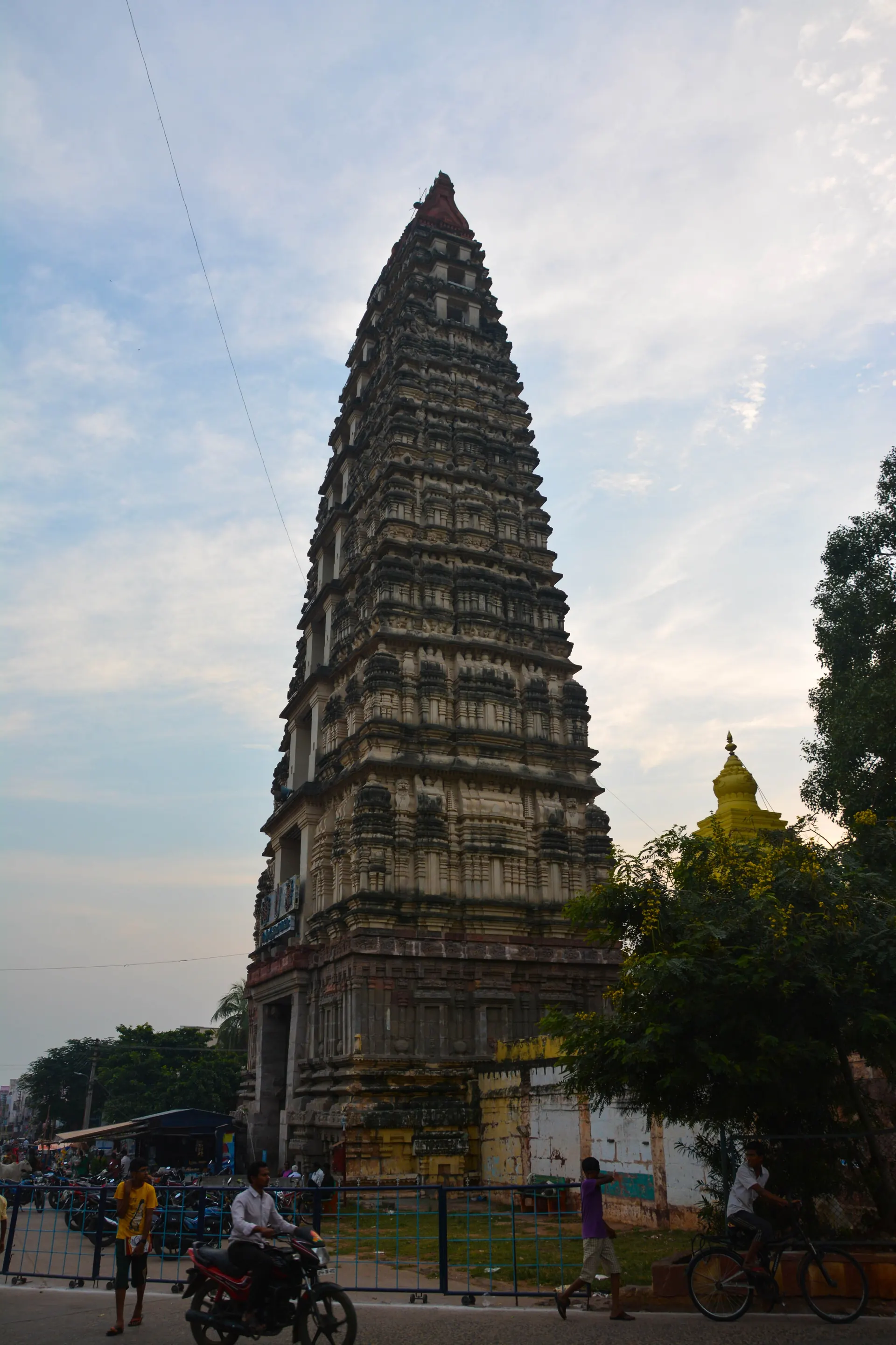 A very tall, narrow stepped temple tower with many storeys of carved stone, rising over a street with motorbikes and people at its foot.