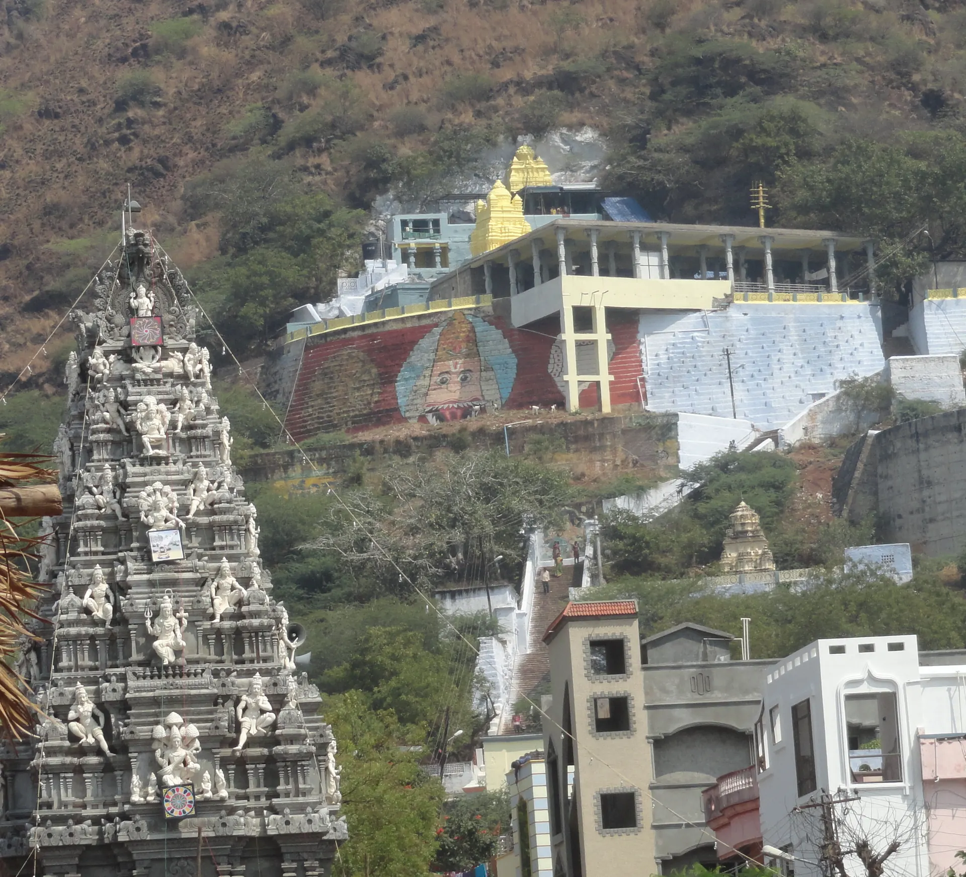 A carved white temple tower in front of a rocky hillside with white temple buildings and steps built up against it.