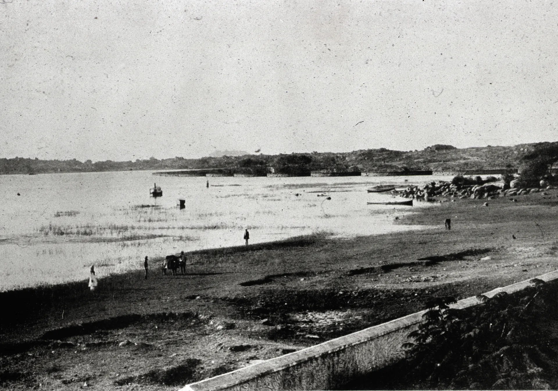 Old black-and-white photograph of a wide shallow lake with a flat shore in front, a few people and a boat on the water, and low hills on the far side.