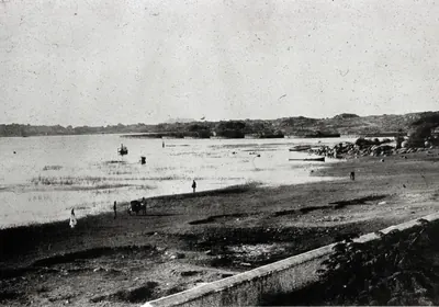 Old black-and-white photograph of a wide shallow lake with a flat shore in front, a few people and a boat on the water, and low hills on the far side.