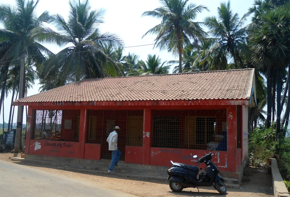 A small single-storeyed house with a red tiled roof and red walls, with a scooter parked in front and coconut palms behind.