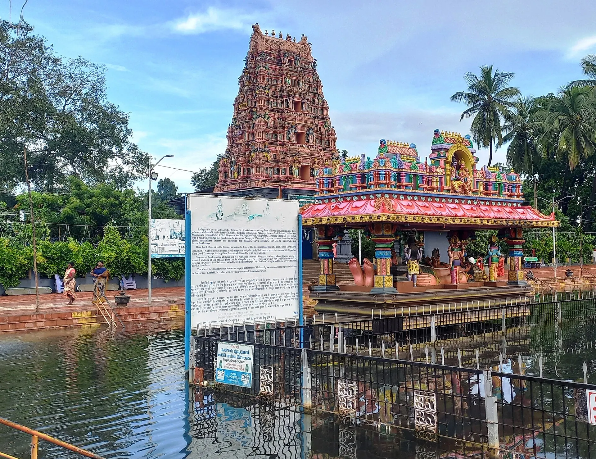 A tall painted temple gateway tower and a colourful shrine on the edge of a large water tank, with railings and a notice board in front and palm trees behind.