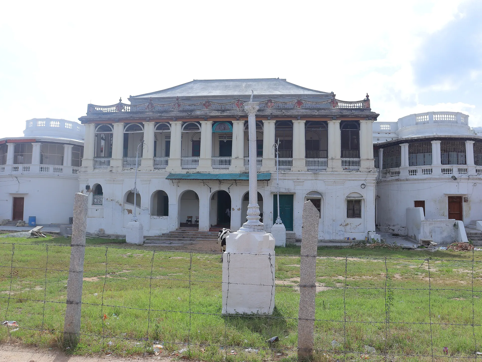 A large white two-storeyed palace with arched windows, pillared balconies and rounded wings on both sides, seen across a grassy yard behind a wire fence.
