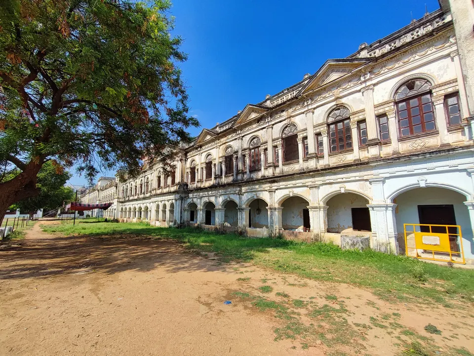 A long cream-coloured two-storeyed palace wing with arched windows and a pillared ground floor, seen across a grassy courtyard under a blue sky.