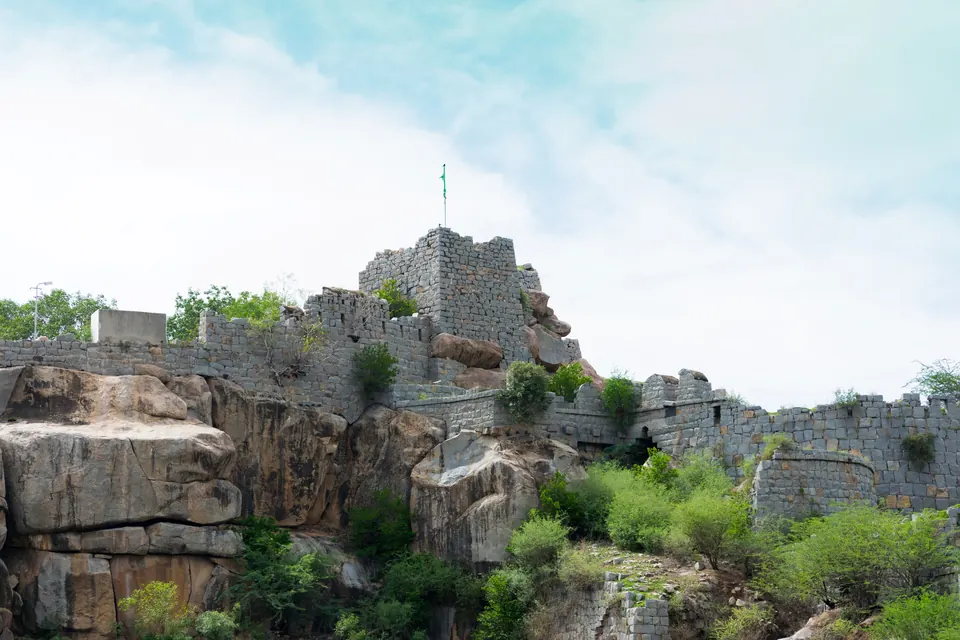 Old grey stone walls and a square bastion built along the top of a hill of large rounded boulders, with bushes below and a pale sky.