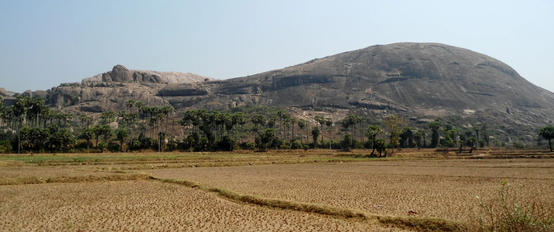 Two long rocky hills with scattered trees rising beyond flat dry fields under a pale sky.