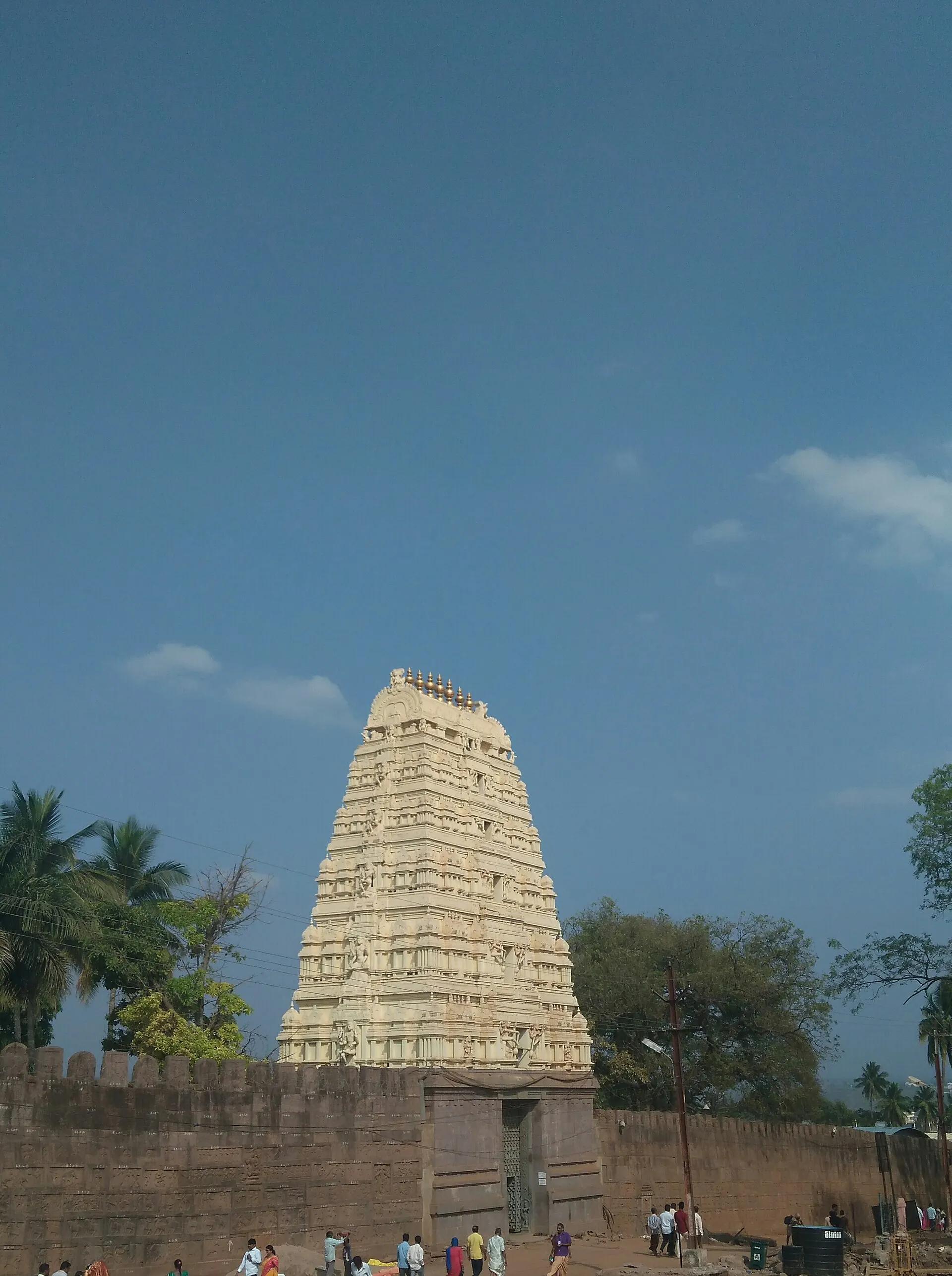 A tall cream-coloured temple gateway tower above a long grey stone wall, with people walking in front under a blue sky.