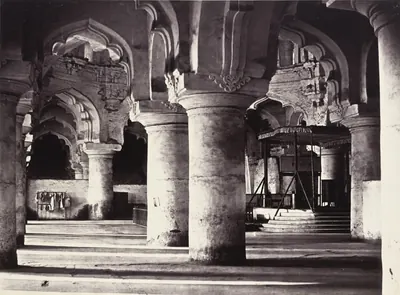 An old sepia photograph of a hall with huge round stone pillars joined by tall pointed, scalloped arches; a raised platform with a canopy stands on the right.