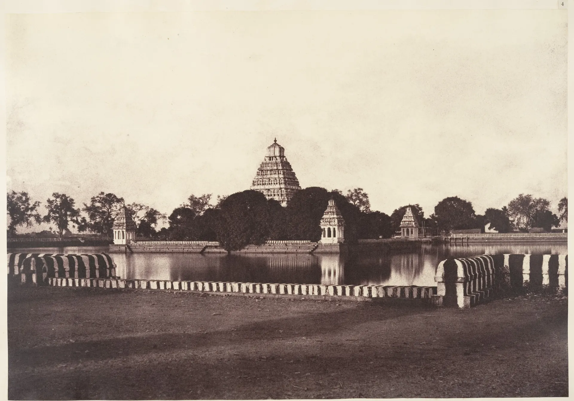 An old photograph of a wide square tank of still water with a striped low wall in front; a temple tower and small pavilions rise from the island in the middle.