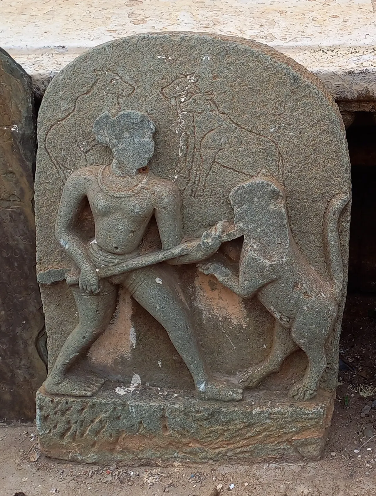 A grey stone slab with a rounded top, carved with a warrior fighting an animal, with worn lines of letters on the stone.