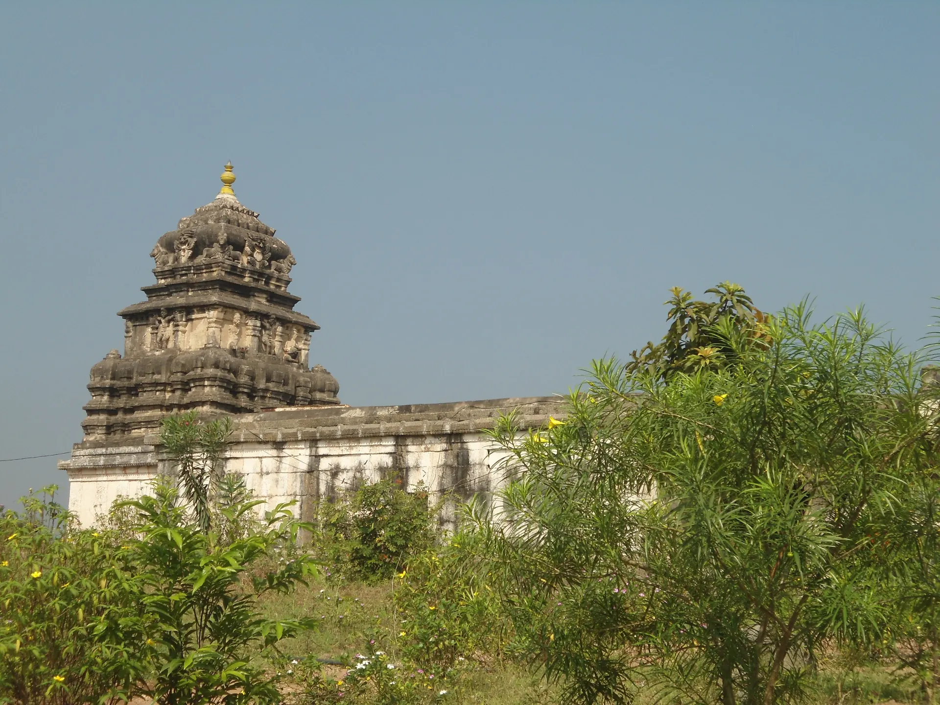 A whitewashed stone temple with a dark stepped tower over the shrine, half hidden by green bushes, under a hazy sky.