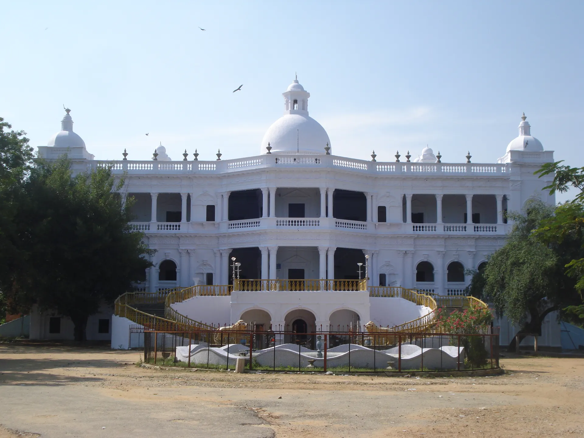 A large white two-storeyed palace with arched verandas, small domes on the roof and a curving double staircase in front of a bare yard.