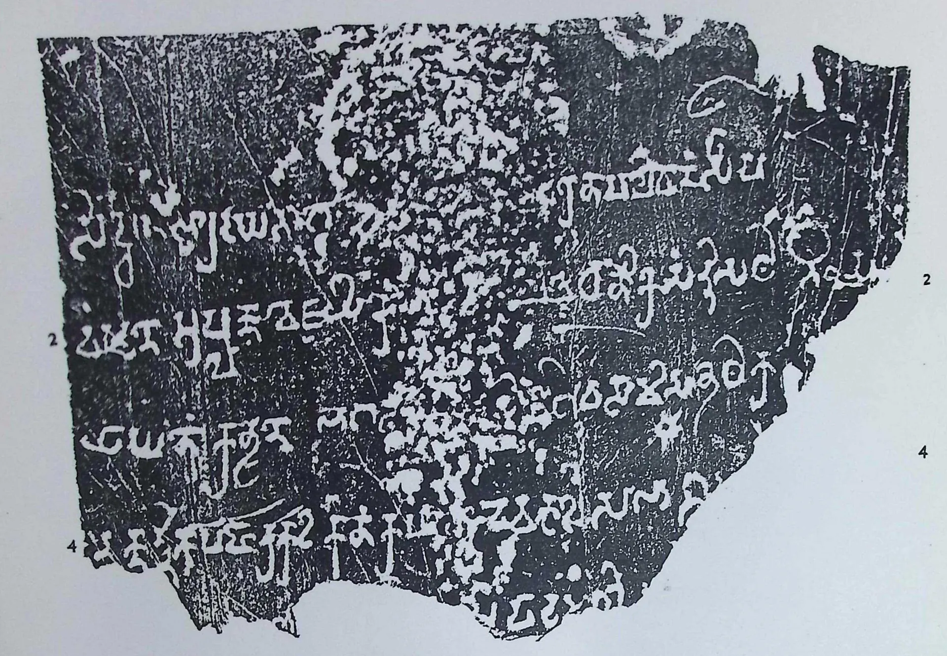 Ink impression of a broken stone slab, irregular outline, with five lines of large Prakrit letters in early southern Brahmi, white on a mottled grey-black ground; line numbers 2 and 4 in the margins.