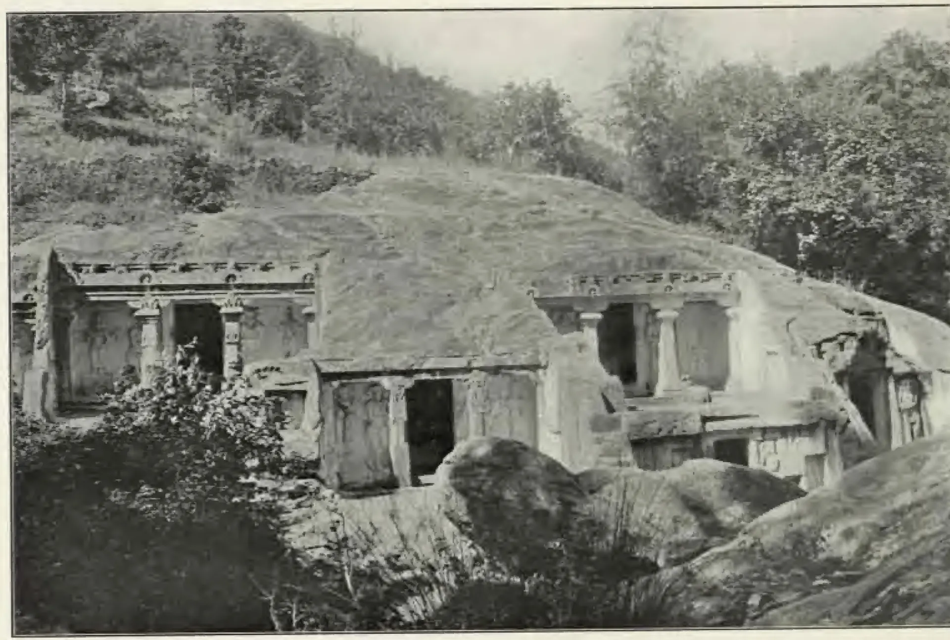 Halftone photograph of a rocky ravine side with several small rock-cut shrines, their pillared fronts cut into the base of a sloping granite outcrop, with trees above and boulders in front.