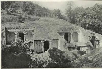 Halftone photograph of a rocky ravine side with several small rock-cut shrines, their pillared fronts cut into the base of a sloping granite outcrop, with trees above and boulders in front.
