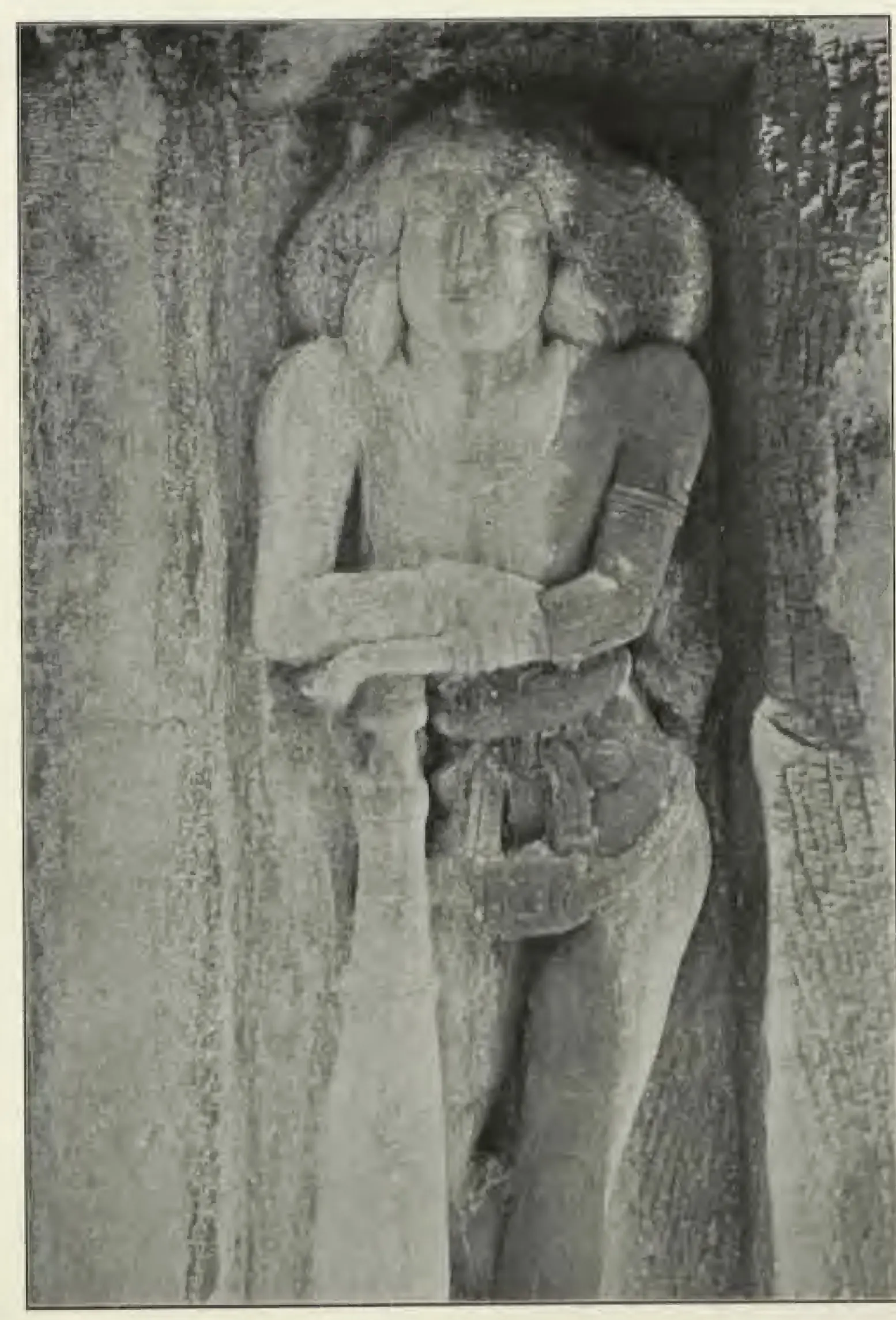 Halftone photograph of a life-size rock-cut door-guardian in a niche, standing with one hand resting on a huge club, the hair spread out in a mass on both sides of the face.