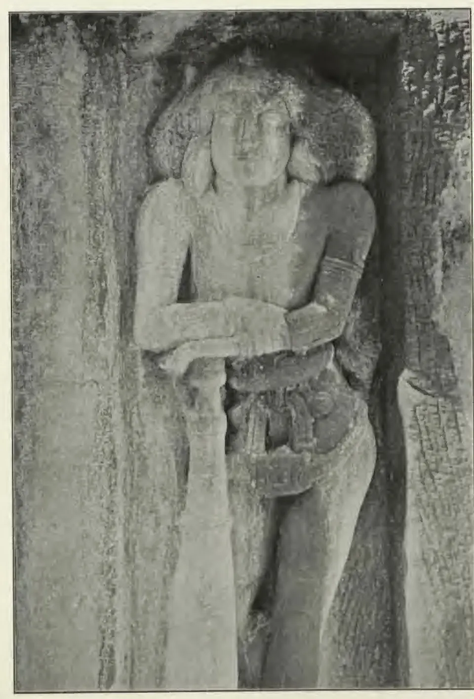 Halftone photograph of a life-size rock-cut door-guardian in a niche, standing with one hand resting on a huge club, the hair spread out in a mass on both sides of the face.