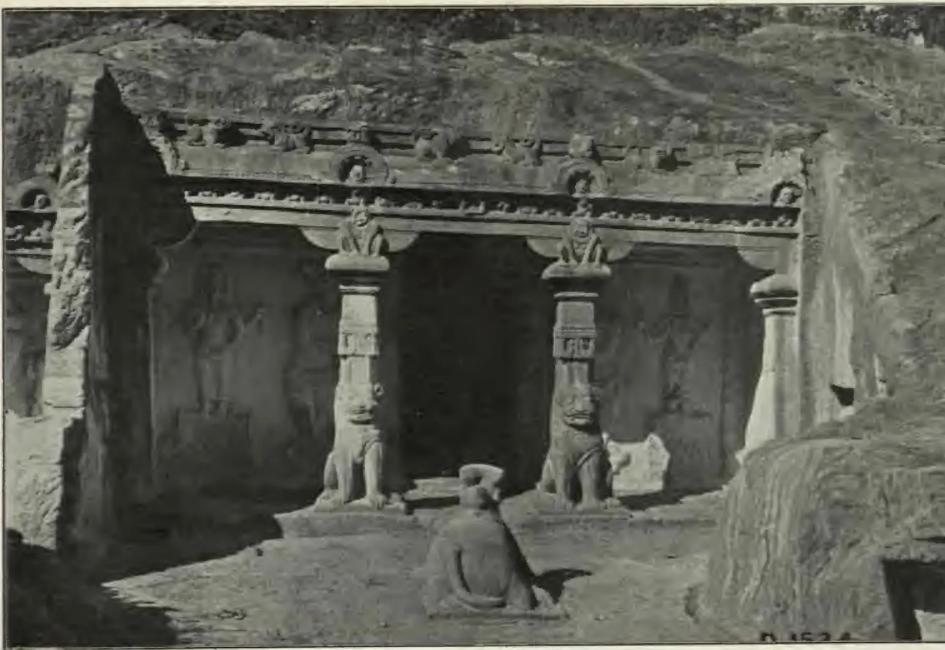 Halftone photograph of a rock-cut shrine front with two pillars carved with seated lions at their bases, a carved cornice above, and a seated stone bull on the terrace in front.