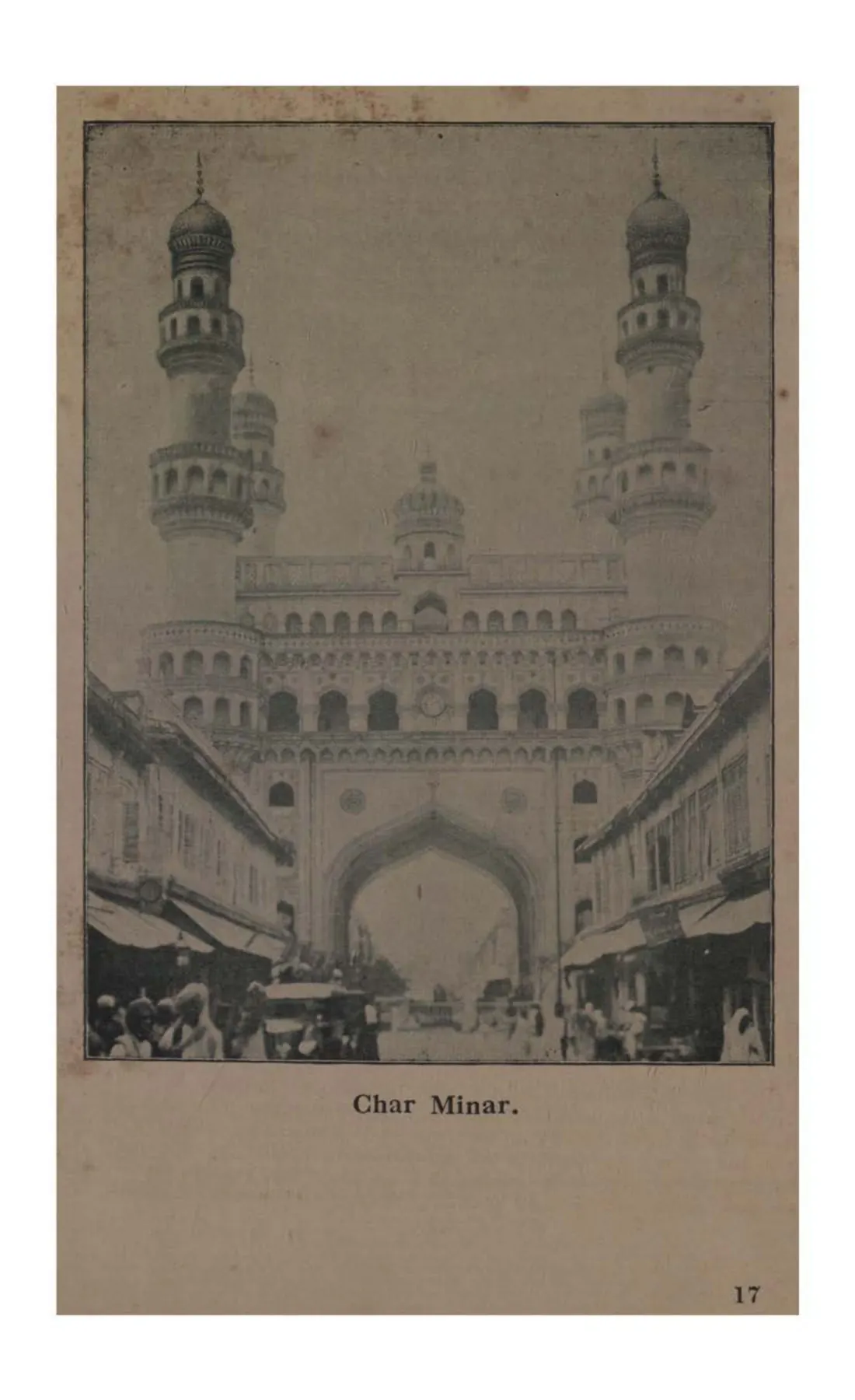 Photograph of the Char Minar seen down a street, its four minarets rising above the arched central block, with shops and people in the foreground.
