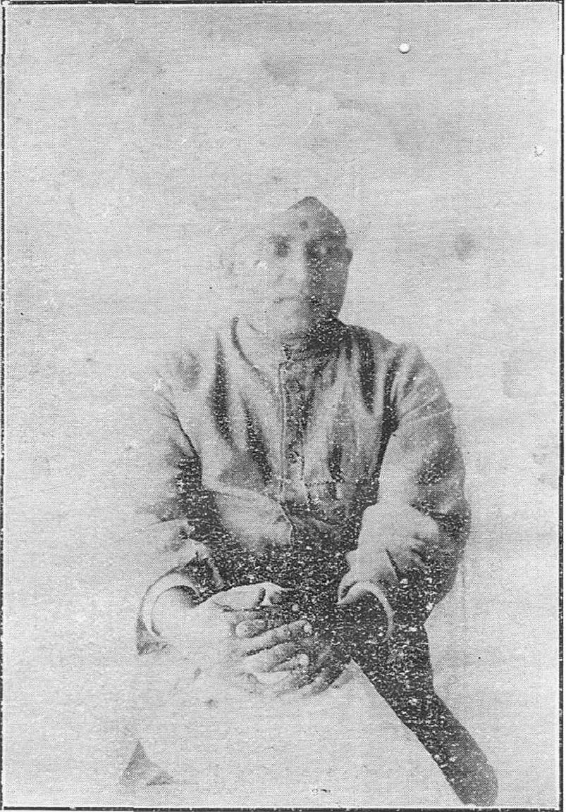 A faded black-and-white half-tone photograph of a seated man in a cap and a long buttoned coat, with his hands folded in his lap.