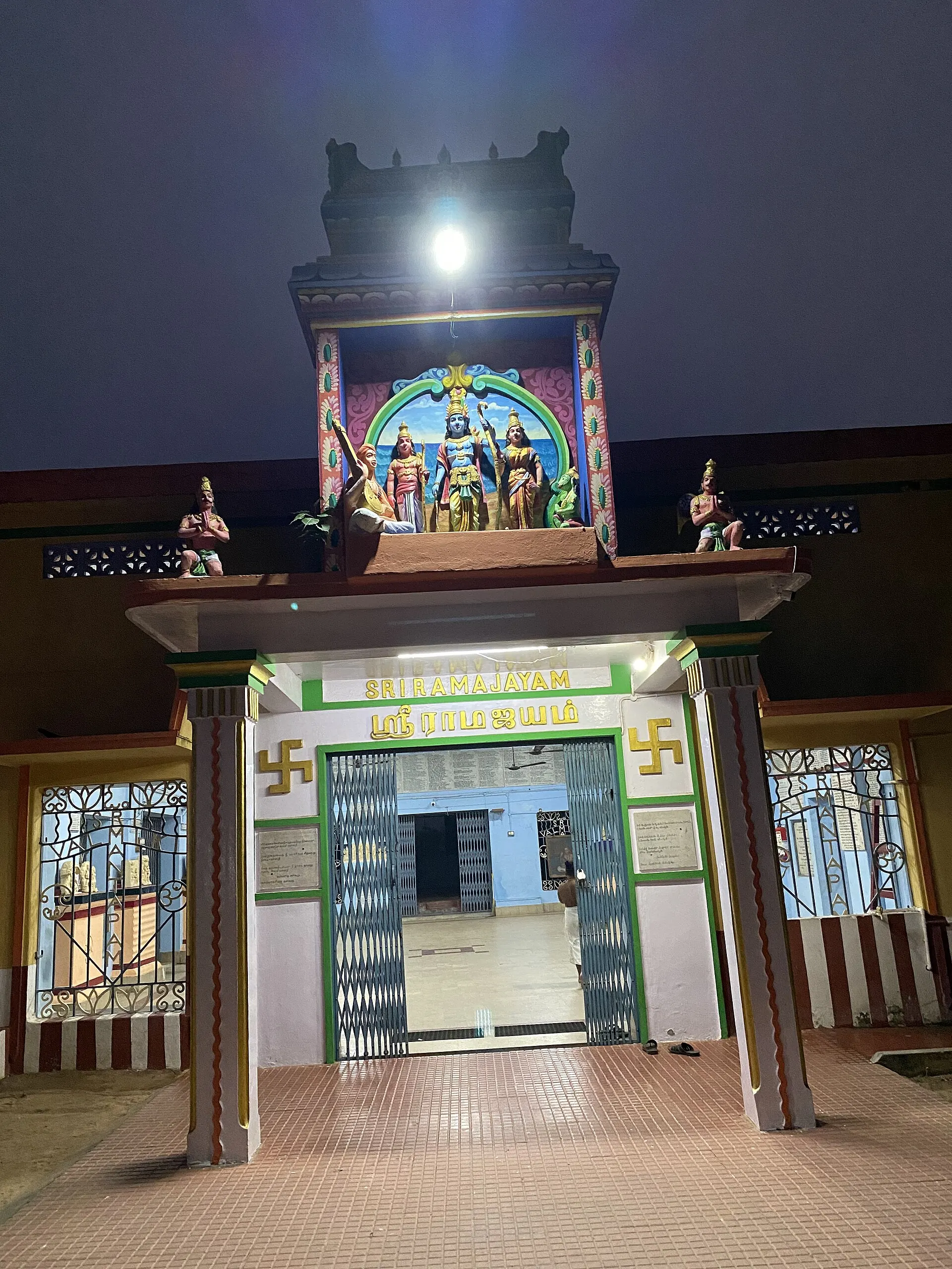 A night photograph of a lit temple gateway with a small painted tower of figures above a doorway with green-and-white name boards, opening onto a paved courtyard.