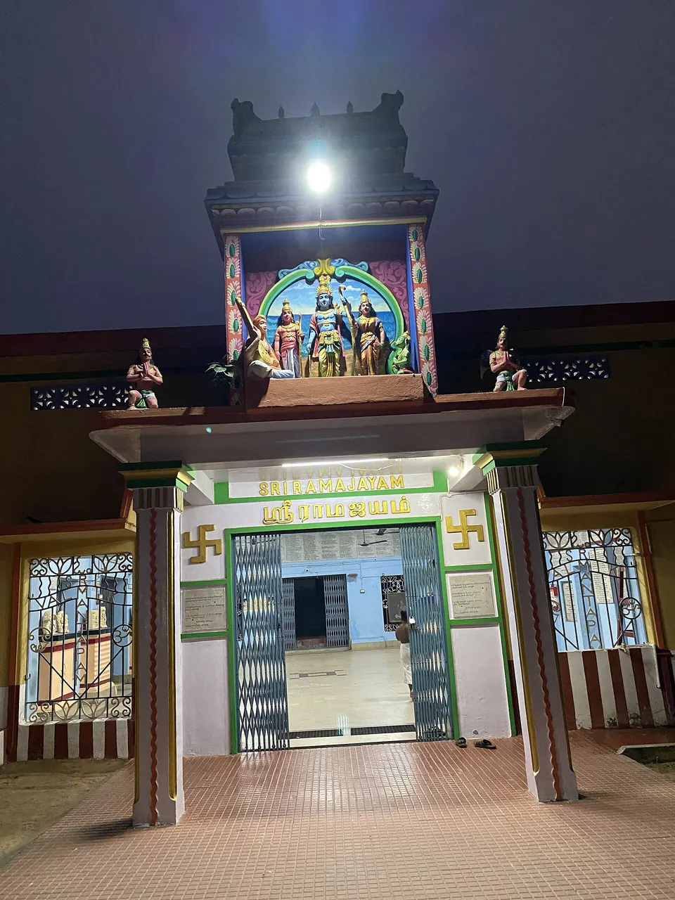 A night photograph of a lit temple gateway with a small painted tower of figures above a doorway with green-and-white name boards, opening onto a paved courtyard.