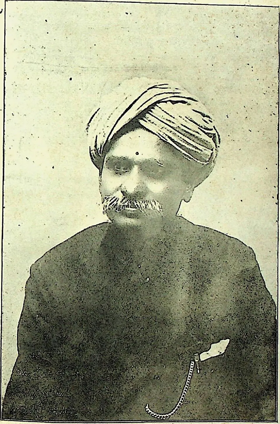 An old half-tone photograph of a man with a white moustache, a striped turban and a dark buttoned coat with a watch chain.
