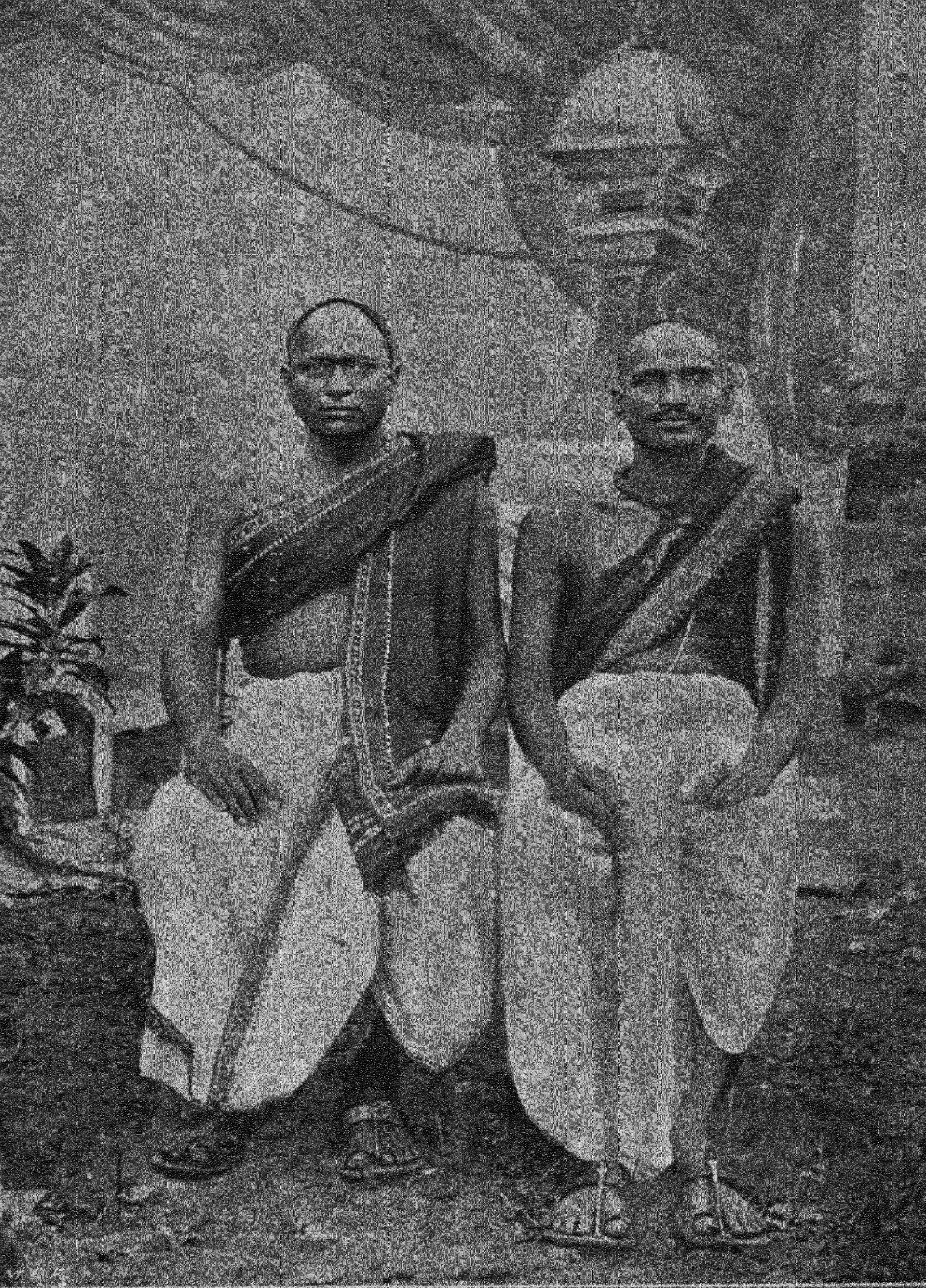 An old half-tone photograph of two seated men with shaven heads, wearing white dhotis and dark upper cloths draped over one shoulder, with a painted backdrop of a tower behind them.