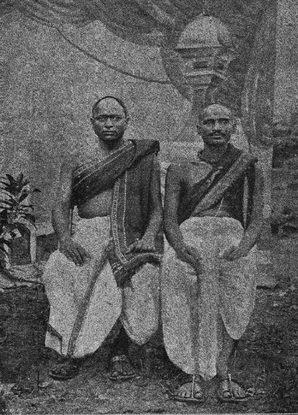 An old half-tone photograph of two seated men with shaven heads, wearing white dhotis and dark upper cloths draped over one shoulder, with a painted backdrop of a tower behind them.