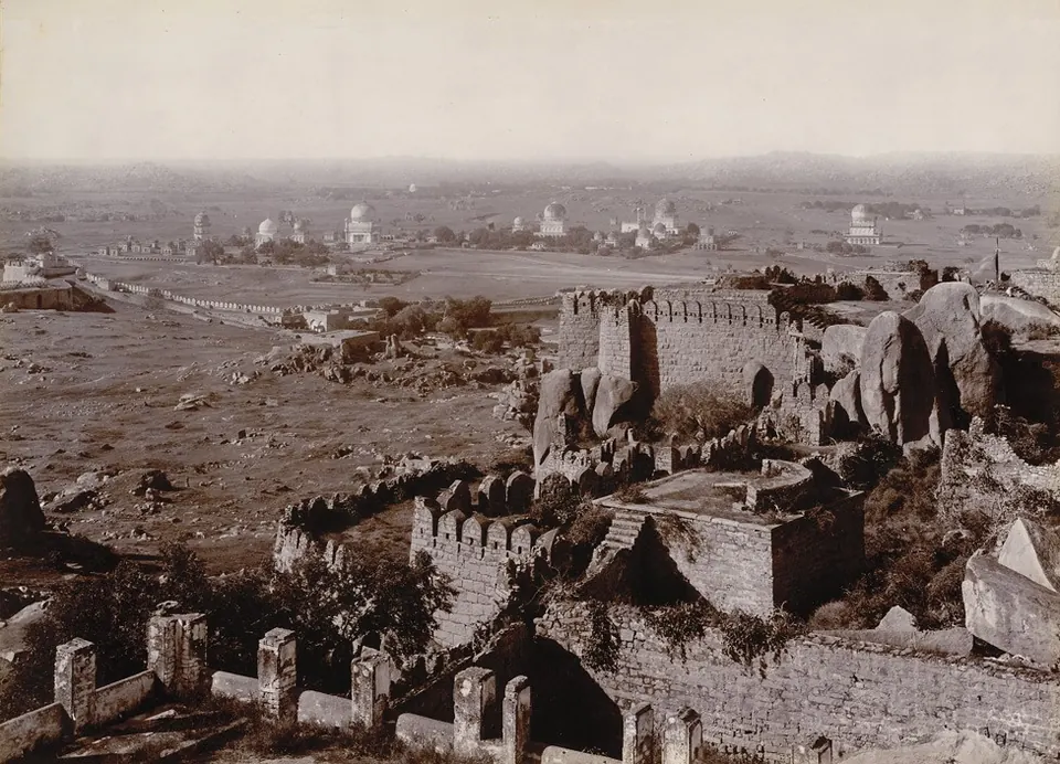 Old photograph looking down from the fort over rocky ground and ruined walls to the domes of the royal tombs on the plain.
