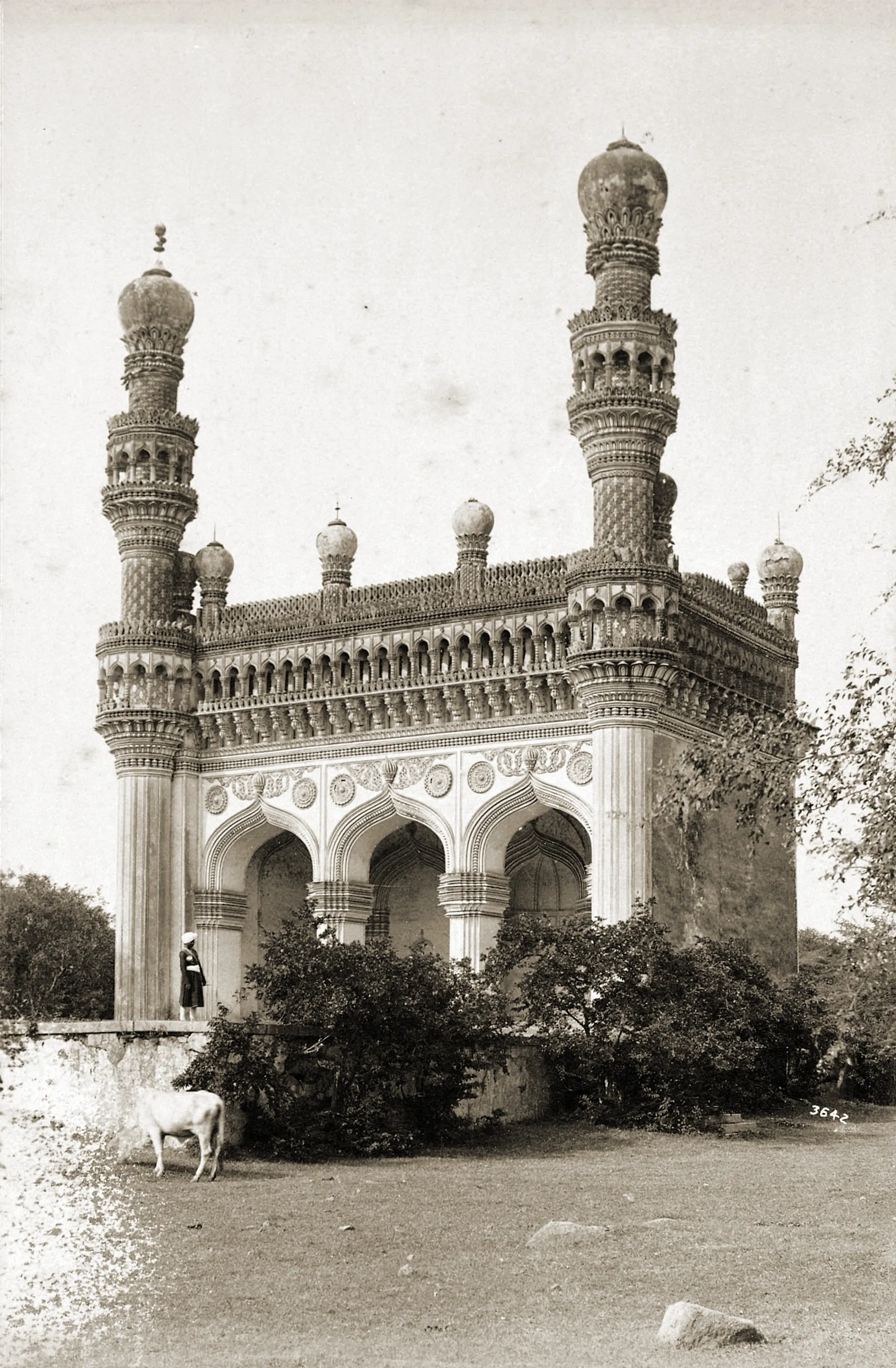 Old photograph of a small mosque with three pointed arches between two tall minarets topped by bulbous domes, trees and bushes around it.