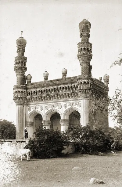 Old photograph of a small mosque with three pointed arches between two tall minarets topped by bulbous domes, trees and bushes around it.