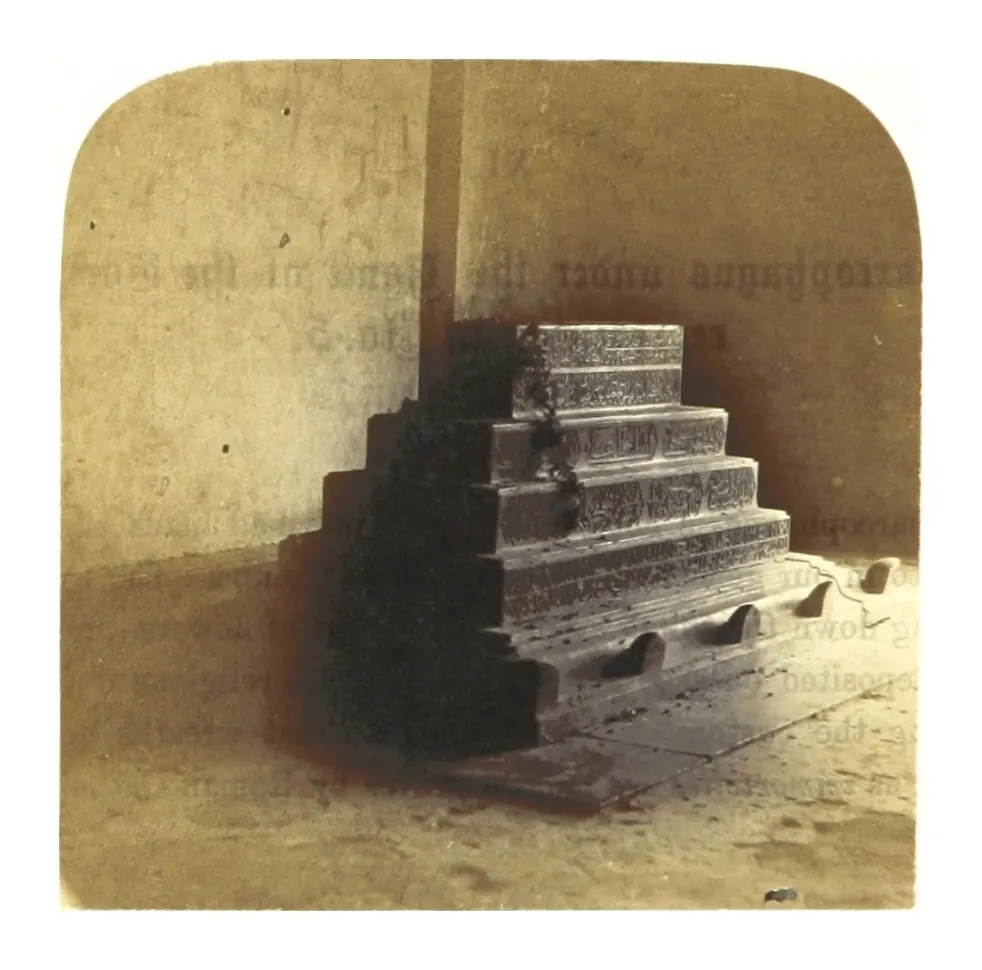 Old photograph inside a tomb chamber: a stepped stone sarcophagus in dim light against a plain wall.