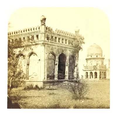 Old photograph of the corner of a square arcaded tomb with a parapet, a smaller domed tomb beyond.