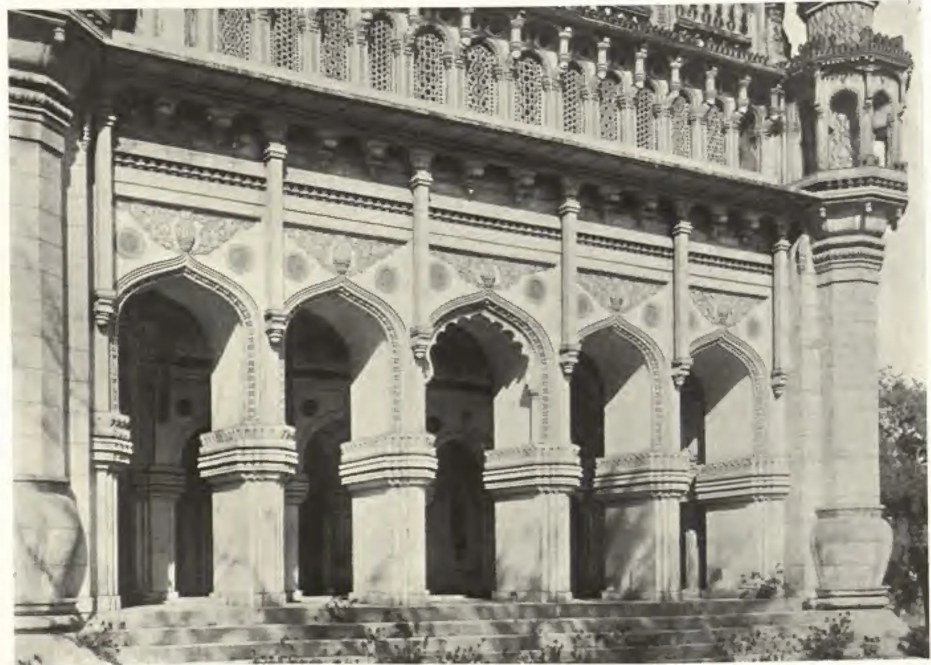 Photograph of the front arcade of the Toli Masjid: pointed arches on heavy piers, a band of carved brackets and pierced screens above, and the base of a minaret at the right.