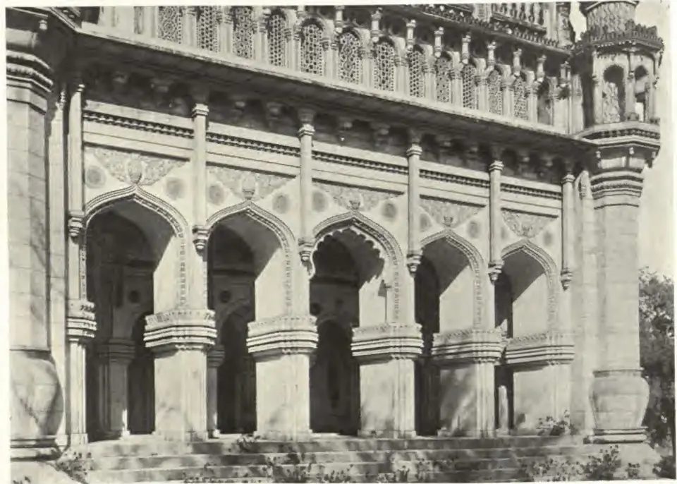 Photograph of the front arcade of the Toli Masjid: pointed arches on heavy piers, a band of carved brackets and pierced screens above, and the base of a minaret at the right.