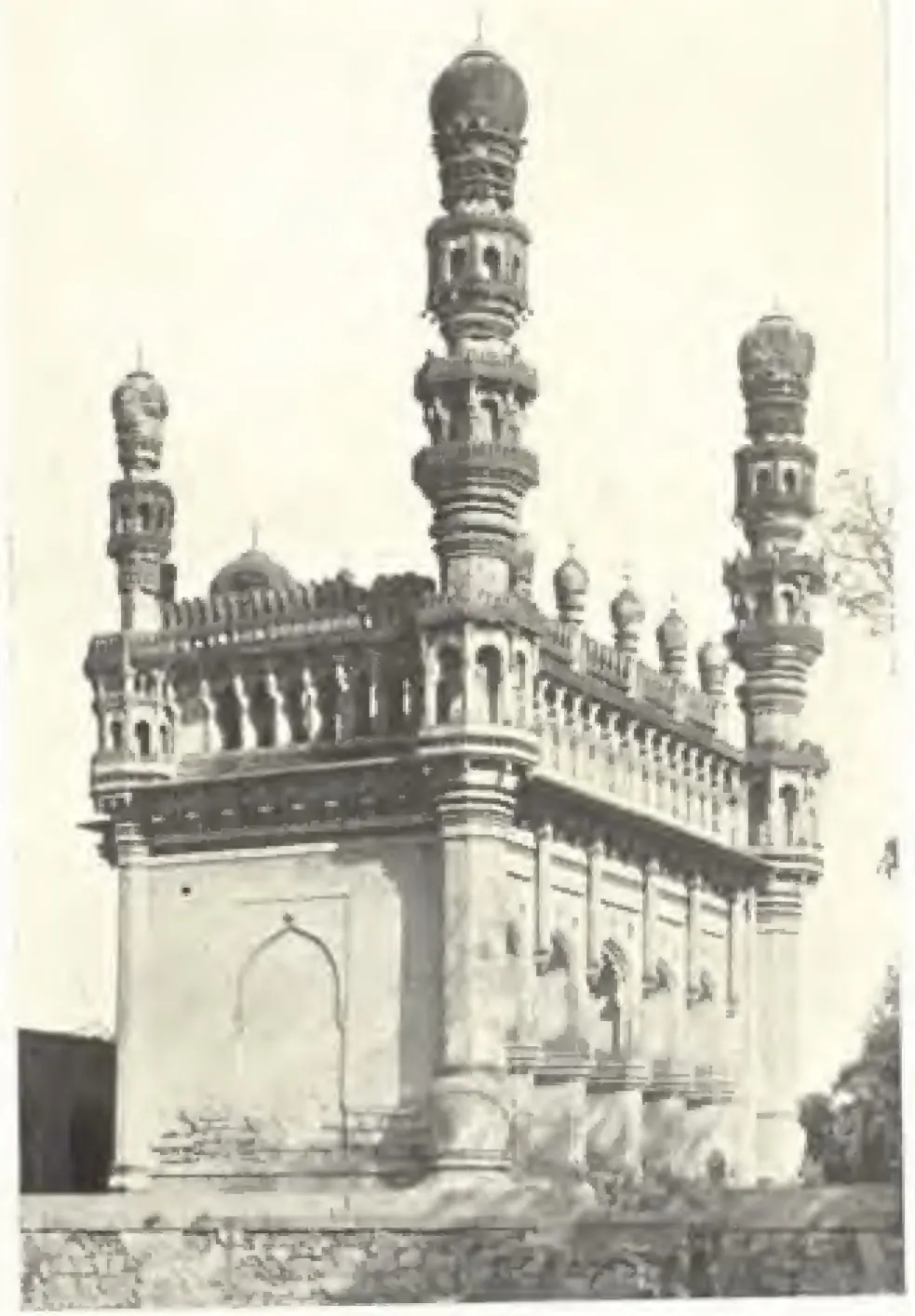 Photograph of the corner of a mosque on a high plinth, its arcaded facade topped by a pierced parapet and small turrets, with two tall many-tiered minarets rising at the corners.