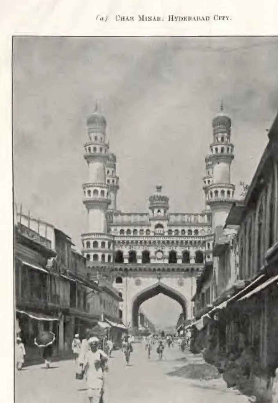 Photograph down a busy street lined with shops towards the Charminar, its great arch and four minarets closing the view, with people walking in the road. Printed sideways.