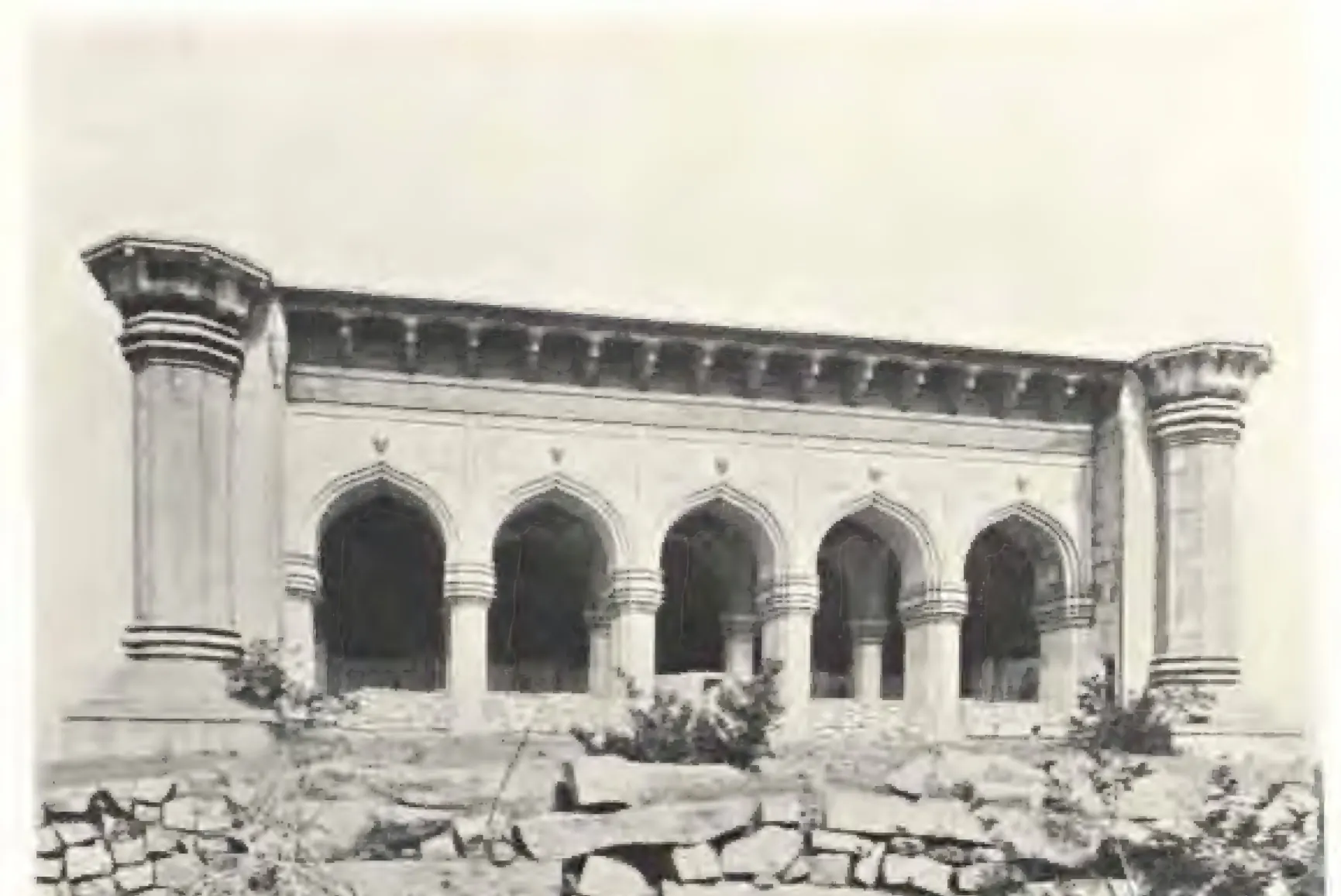 Photograph of the arcaded front of a mosque on a rocky terrace: five pointed arches on square piers under a deep eave, with the stumps of two minarets at the ends.