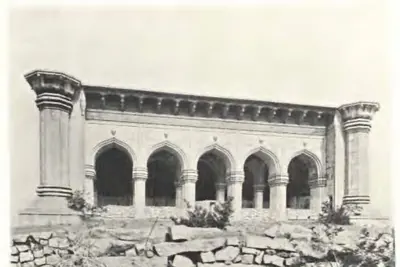 Photograph of the arcaded front of a mosque on a rocky terrace: five pointed arches on square piers under a deep eave, with the stumps of two minarets at the ends.