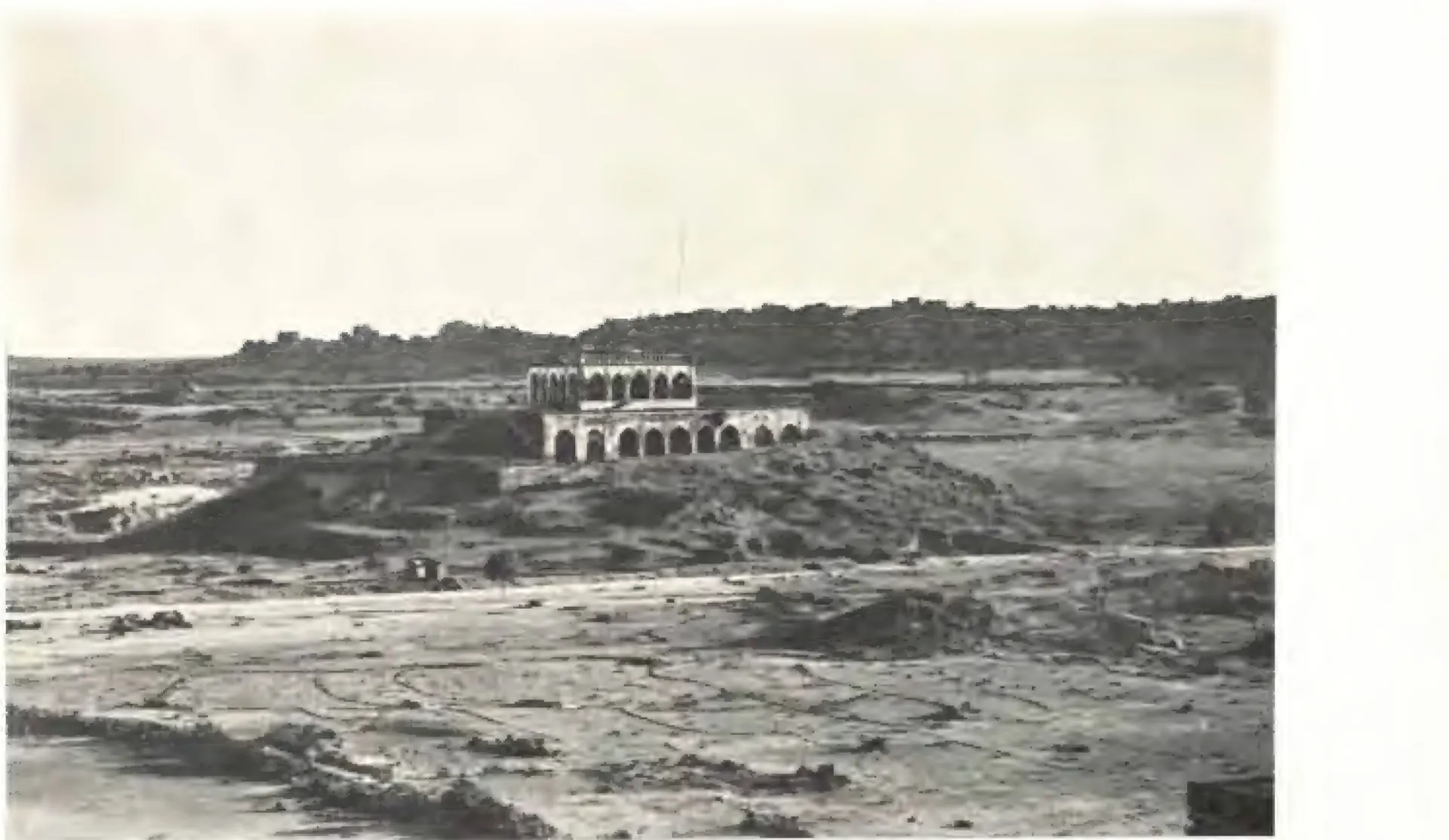 Photograph across stony open ground to a low rocky hill crowned by a long arcaded pavilion on stepped terraces.