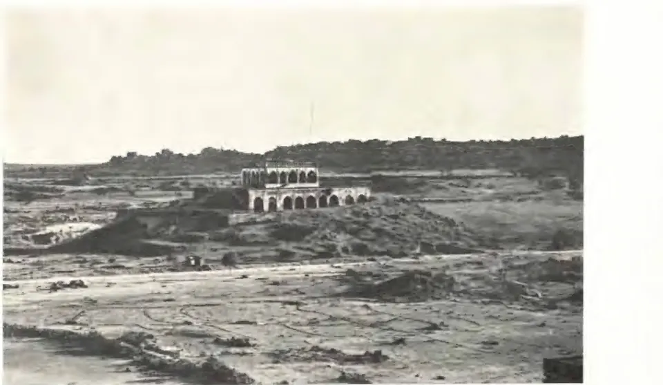 Photograph across stony open ground to a low rocky hill crowned by a long arcaded pavilion on stepped terraces.