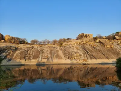 Colour photograph of a bare sloping granite hill with ruined walls and a small building on its crest, reflected in a still pond at its foot.