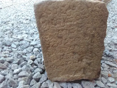 Colour photograph of a weathered upright stone slab carved with rows of worn letters, standing on a bed of gravel.
