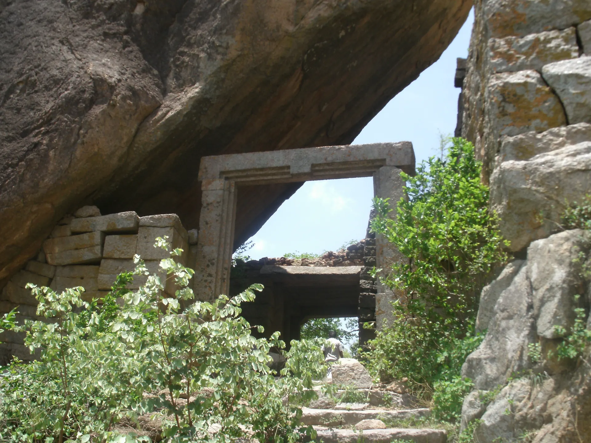 Colour photograph of a stone doorway with a heavy lintel built beneath a huge overhanging granite boulder, with steps and shrubs in front and walls of rough blocks beyond.