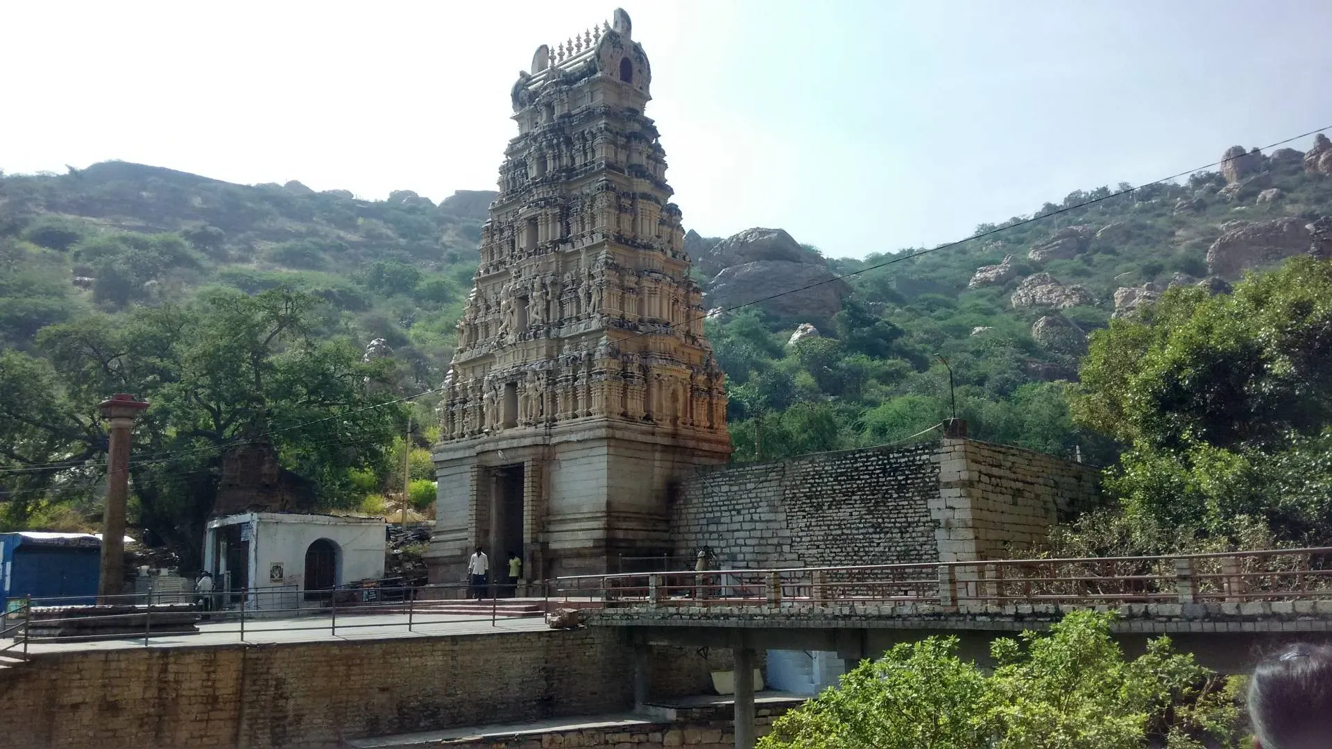 Colour photograph of a tall stepped gateway tower and temple buildings at the foot of a wooded rocky hill, with a railed walkway in front.