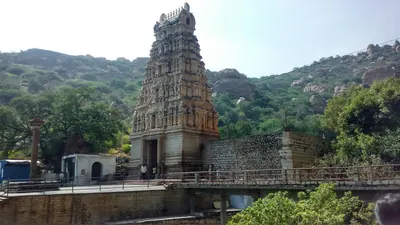 Colour photograph of a tall stepped gateway tower and temple buildings at the foot of a wooded rocky hill, with a railed walkway in front.