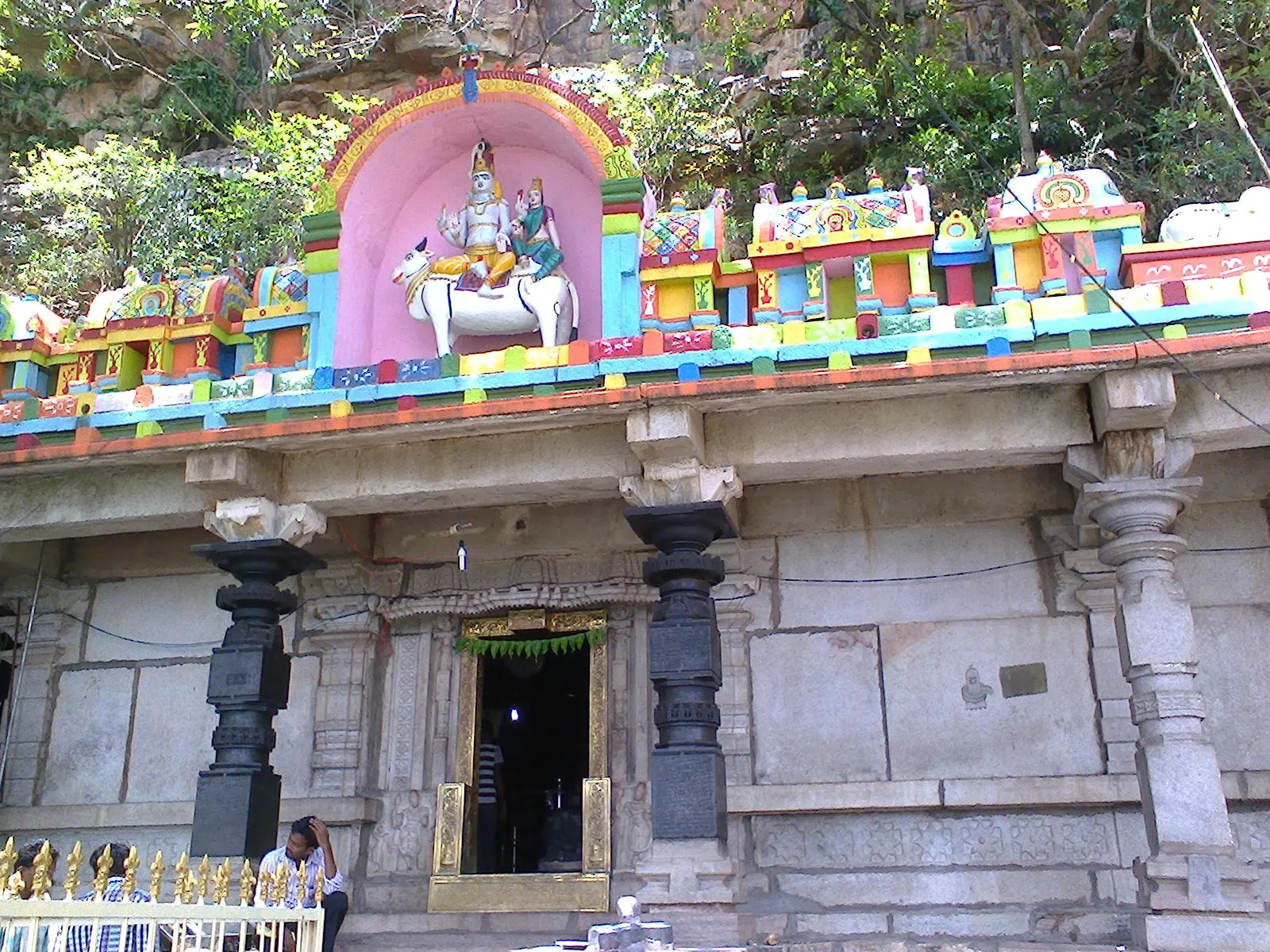 Colour photograph of a stone temple front with black carved pillars and a doorway, below a brightly painted parapet with a stucco group of Siva and Parvati on the bull Nandi.