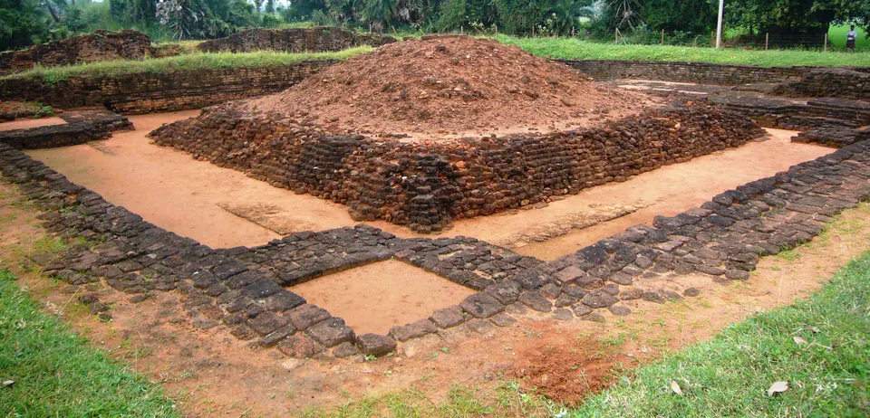 An excavated square brick platform with a mound of loose rubble on top, inside a sunken trench lined with later brick walls, with grass and trees around.